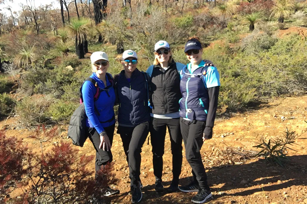 The four Cape to Cape walkers arm in arm on a bush track in the south west, wearing caps and hiking packs